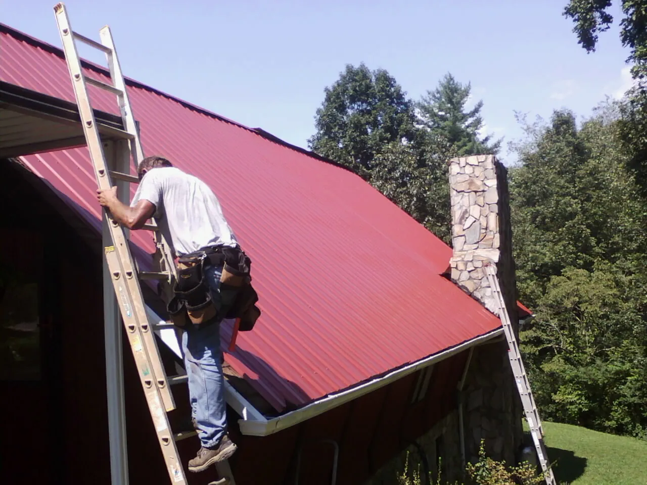 Roofer on a ladder beside a red metal roof and stone chimney — Soffit & Fascia Repair by Roofing Brothers in White Castle